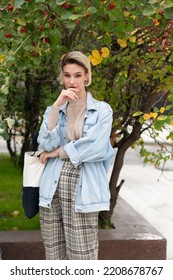 Portrait Of A Young Beautiful Woman In A Denim Jacket Outside, Stands Near A Green Bush