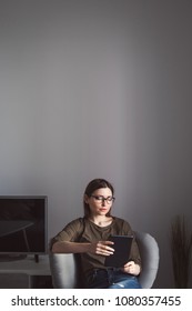 Portrait Of Young Beautiful Woman In Glasses Sitting On A Chair With Ipad Laptop In Her Hands And Reading.