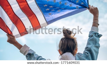 Portrait of a young beautiful woman in glasses and jeans with a waving American flag in her hands, background of blue sky, concept of patriotism, demonstration, protest.