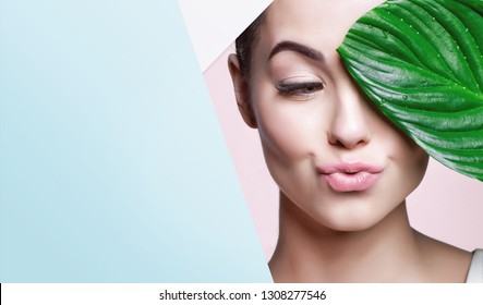 Portrait of young beautiful woman with healthy glow perfect smooth skin holds green tropical leaf, look into the hole of colored paper. Model with natural nude make up. Fashion, beauty, skincare.