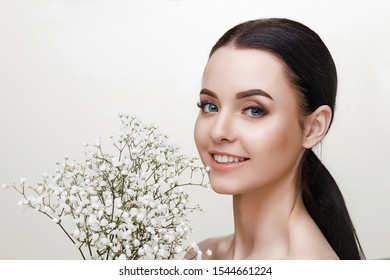 Portrait of young beautiful woman with healthy glow perfect smooth skin and blue eyes holds flowers. Model with natural nude make up. Gray background.