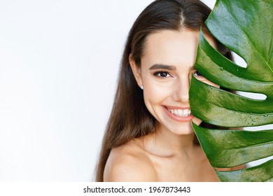 Portrait of young beautiful woman with healthy glow skin holds green tropical leaf and covers part of her face. Model with gentle makeup. Personal care, cosmetology concept.