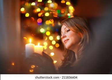 A Portrait Of A Young Beautiful Woman Holding A Candle And Looking At It With Twinkling Fairy Lights As A Background. Christmas.