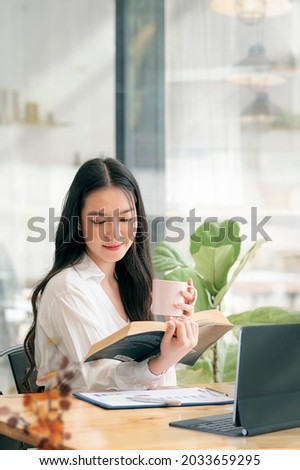 Portrait of young beautiful woman holding mug and reading a book while relaxing during work at home. Vertical view.