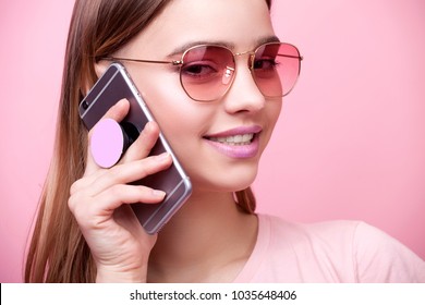 Portrait of young beautiful woman isolated on pink background in t-shirt of the same color. An attractive smiling girl speaks on mobile phone on which is attached modern holder for phone pop socket