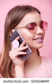 Portrait of young beautiful woman isolated on pink background in t-shirt of the same color. An attractive smiling girl speaks on mobile phone on which is attached modern holder for phone pop socket
