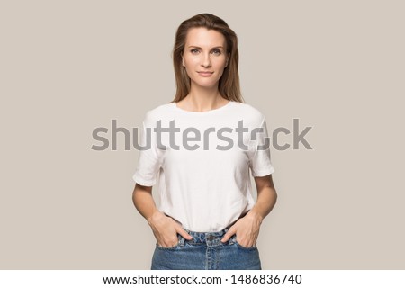 Portrait of young beautiful woman in jeans and T-shirt hold hands in pockets look at camera pose for picture, pretty millennial Caucasian female model with long hair isolated on grey studio background
