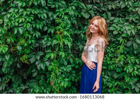 Portrait of a young beautiful woman with make up and curly blond hair, standing next to the wall of wild grape leaves. Copy space.