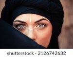 Portrait of a young beautiful woman with makeup and white face art ornament in tribal or middle eastern style dressed in a black hijab. Ethnic image Close-up on the background of a sandy wall.