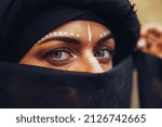 Portrait of a young beautiful woman with makeup and white face art ornament in tribal or middle eastern style dressed in a black hijab. Ethnic image Close-up on the background of a sandy wall.