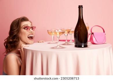 Portrait of young beautiful woman peeking out table served with champagne against pink studio background. Excitement and happiness. Concept of emotions, celebration, party, fashion