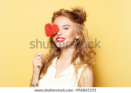 Portrait of young beautiful woman with pink lipstick, curly long hair and playful hairstyle. Smiling. Holding and licking a heart shaped lollipop. Dark yellow background. Valentines day. 