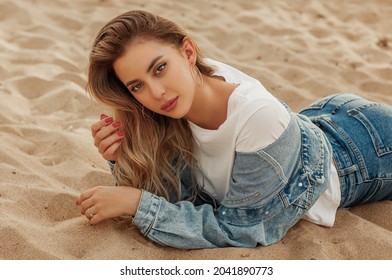 Portrait Of Young Beautiful Woman Posing On Sand, Playing With Her Long Hair. Model Wearing Stylish Blue Denim Jacket, Top, Jeans. Sunset Light. Female Fashion Concept.