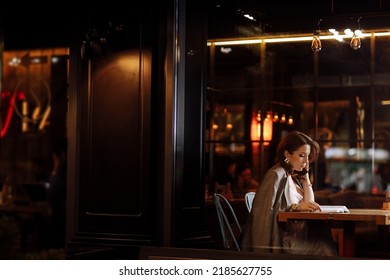 portrait of young beautiful woman is reading paper book in cafe in dusk. girl is online study, remote work