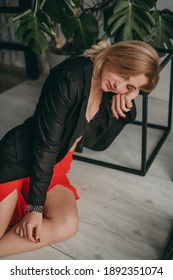 Portrait Of Young Beautiful Woman In Red Pants With Strut, Top, Bra And Black Jacket At Home Near A Large Mirror, Metal Cube And Triangle, Flowerpot. Scandinavian Interior. White Walls, Cozy, Fashion.