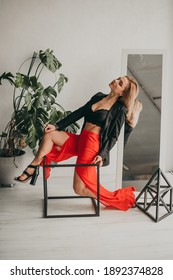 Portrait Of Young Beautiful Woman In Red Pants With Strut, Top, Bra And Black Jacket At Home Near A Large Mirror, Metal Cube And Triangle, Flowerpot. Scandinavian Interior. White Walls, Cozy, Fashion.