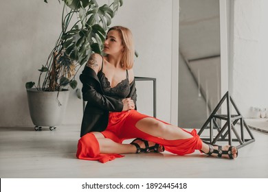 Portrait Of Young Beautiful Woman In Red Pants With Strut, Top, Bra And Black Jacket At Home Near A Large Mirror, Metal Cube And Triangle, Flowerpot. Scandinavian Interior. White Walls, Cozy, Fashion.