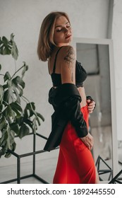 Portrait Of Young Beautiful Woman In Red Pants With Strut, Top, Bra And Black Jacket At Home Near A Large Mirror, Metal Cube And Triangle, Flowerpot. Scandinavian Interior. White Walls, Cozy, Fashion.