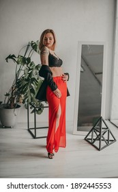 Portrait Of Young Beautiful Woman In Red Pants With Strut, Top, Bra And Black Jacket At Home Near A Large Mirror, Metal Cube And Triangle, Flowerpot. Scandinavian Interior. White Walls, Cozy, Fashion.