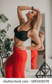 Portrait Of Young Beautiful Woman In Red Pants With Strut, Black Top, Bra At Home Near A Large Mirror, Metal Cube And Triangle, Flowerpot. Scandinavian Interior. White Walls, Cozy, Fashion.