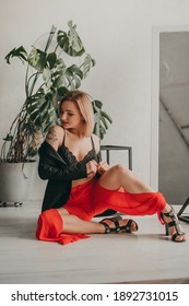 Portrait Of Young Beautiful Woman In Red Pants With Strut, Top, Bra And Black Jacket At Home Near A Large Mirror, Metal Cube And Triangle, Flowerpot. Scandinavian Interior. White Walls, Cozy, Fashion.