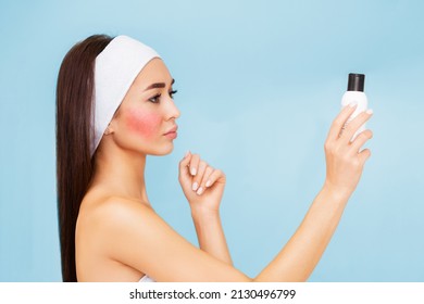 Portrait of a young beautiful woman with rosacea on her cheek, holding a bottle of cosmetics in her hands. The concept of caring for sensitive skin.