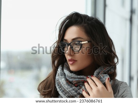 Portrait of young beautiful woman in scarf, warming hands, pensive look away, view from window. Brunette lady in glasses, mysterious look. Beauty concept
