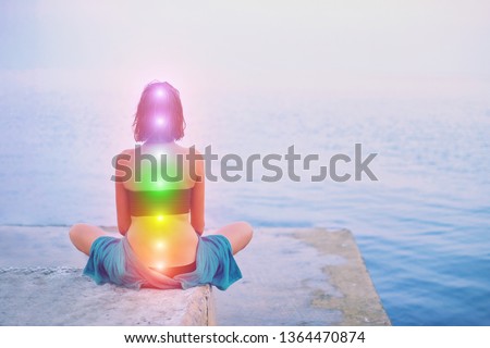 Portrait of young beautiful woman sits in a pose of half lotus practicing yoga meditation at blue summer sea environment glowing all seven chakra Kundalini energy exercise Back view Inner mind consept