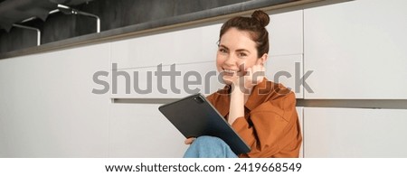 Portrait of young beautiful woman sitting on kitchen floor with digital tablet, browsing news feed, social media app on gadget, smiling and looking happy.