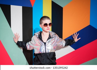 Portrait of young beautiful woman in sunglasses with short hair in a silver jacket on bright geometric background, her hands touch the wall. Fashion concept 