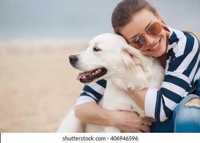 Portrait Of Young Beautiful Woman In Sunglasses Sitting On Sand Beach Hugging Golden Retriever Dog. Girl With Dog By Sea. Happiness And Friendship. Pet And Woman.woman Playing With Dog On Sea Shore