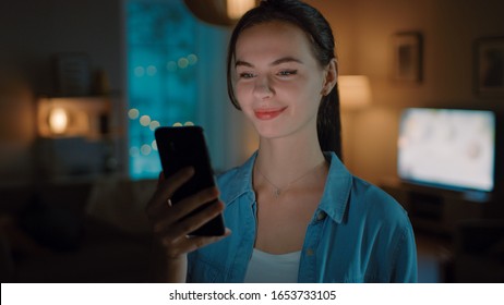 Portrait of Young Beautiful Woman Using Smartphone, Browsing in Internet, Checking Social Networks and Using Smart Home Application while Standing At Home. Cozy Evening.