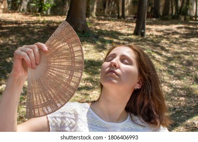 Portrait Of Young Beautiful Woman  Using Hand Fan In Very Hot Summer Day Outdoors. Lady Cooling Herself. Suffering From High Temperature Outside.