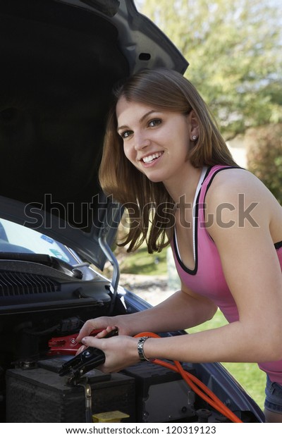 Portrait of a young beautiful woman using jumper\
cables to charge a car\
battery