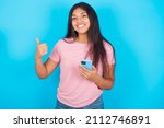 Portrait of Young beautiful woman wearing pink T-shirt against blue background using and texting with smartphone  happy with big smile doing ok sign, thumb up with fingers, excellent sign