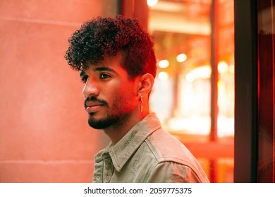 Portrait Of Young Biracial Curly Haired Man With Orange Neon Light Reflection On Face. Looking To The Side. Millennial With Beard And Earring. Urban Style. Horizontal Image.