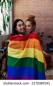 A Portrait Of A Young Bisexual Couple Are Caped With A Pride Flag In Their Living Room And Looking At The Camera