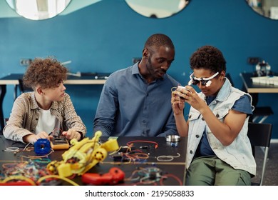 Portrait Of Young Black Boy Building Robots In Engineering Class With Male Teacher Helping