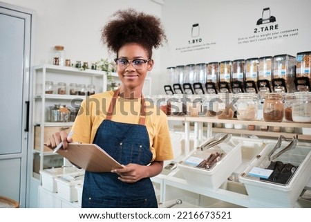 Portrait of young Black female shopkeeper smiling and looking at camera arranging natural products at refill store, zero-waste grocery, and plastic-free, environment-friendly with reusable containers.