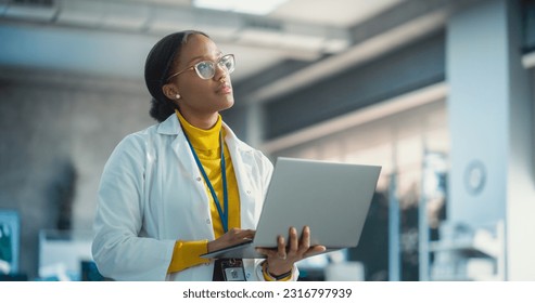 Portrait of Young Black Female Specialist in a Lab Coat Using Laptop Computer. Professional and Successful Woman Working as an Engineer, Thinking and Developing High Technology Projects In Startup - Powered by Shutterstock - Get 15% off with code: PIKWIZARD15