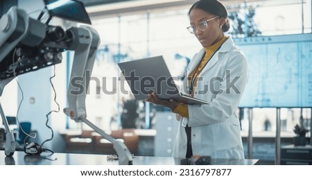 Portrait of Young Black Female Specialist in Lab Coat Using Laptop Computer to Test an AI Robotic Prototype. Professional, Successful Woman Working as an Engineer in High Tech Company Startup
