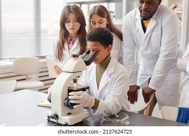 Portrait Of Young Black Girl Looking Into Microscope With While Doing Experiments With Group Of Children In School Chemistry Lab