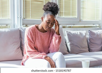 Portrait of a young black girl sitting on the couch at home with a headache and back pain. Beautiful woman suffering from chronic daily headaches. Sad woman holding her head because sinus pain