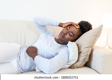 Portrait Of A Young Black Girl Sitting On The Couch At Home With A Headache And Stomach Pain. Beautiful Woman Suffering From Chronic Daily Headaches. Sad Woman Holding Her Head Because Sinus Pain