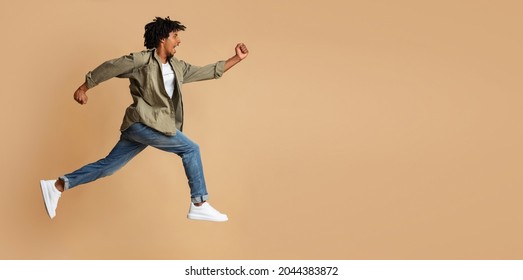 Portrait Of Young Black Guy Running Fast Over Beige Studio Background, In Motion Side View Shot Of Excited African American Man Jump In Air, Hurry Up To Sales Or Discounts, Panorama With Copy Space