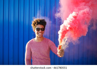 Portrait Of A Young Black Hipster Man Smiling And Holding A Colorful Pink Smoke Bomb Over A Blue Color Background. Youth And Lifestyle Concept