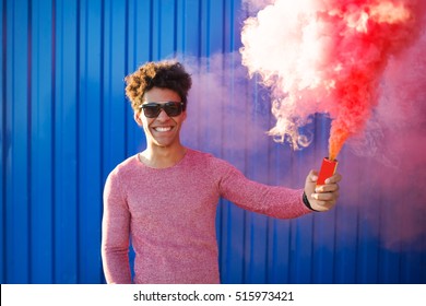 Portrait Of A Young Black Hipster Man Smiling And Holding A Colorful Pink Smoke Bomb Over A Blue Color Background.