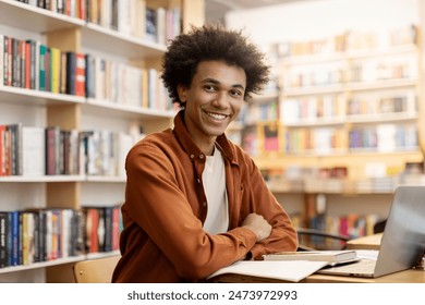 Portrait of young Black male student sitting in university library at table with a laptop computer ad smiling at camera - Powered by Shutterstock