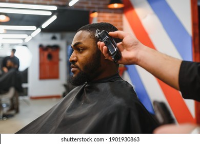 Portrait Of Young Black Man Being Trimmed With Professional Electric Clipper Machine In Barbershop.Male Beauty Treatment Concept. Young African Guy Getting New Haircut In Barber Salon