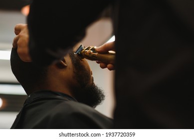 Portrait Of Young Black Man Being Trimmed With Professional Electric Clipper Machine In Barbershop.Male Beauty Treatment Concept. Young African Guy Getting New Haircut In Barber Salon